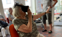 Eine ältere Dame sitzt auf einem Stuhl und trägt eine VR-Brille. Sie greift in die Luft, so als könnte sie dort etwas aus der virtuellen Welt anfassen.