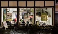 Das Bild zeigt das stadtLABOR Soest von außen. Es ist bereits Abend, aber durch die großen bodentiefen Fenster scheint Licht auf die Straße und reflektiert sich in auf dem regennassen Boden. Drinnen sind viele Menschen zu sehen, die in eine Diskussion vertieft sind.