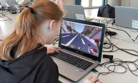 Ein Mädchen sitzt an einem Laptop und programmiert bei Minecraft. Im Hintergrund sind weitere Kinder an Laptops zu sehen.