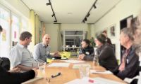 Eine Gruppe von Menschen sitzt gemeinsam an einem langen Tisch. Alle sind in ein Gespräch vertieft, es wird diskutiert und gestikuliert.