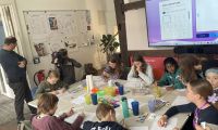 Kinder am Tisch, die malen und ein Fernsehteam im Hintergrund