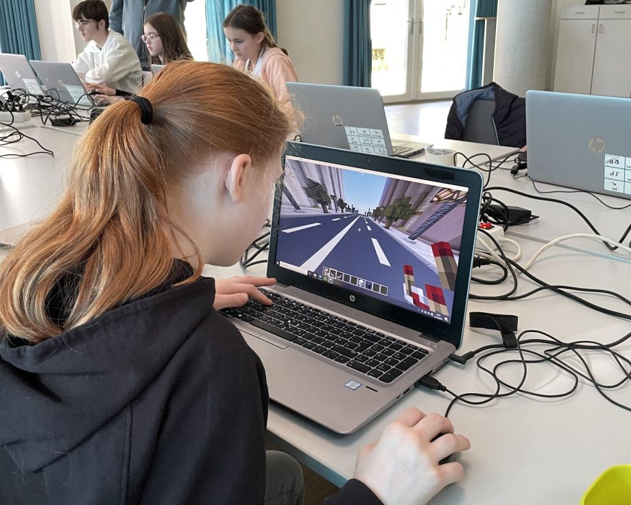 Ein Mädchen sitzt an einem Laptop und programmiert bei Minecraft. Im Hintergrund sind weitere Kinder an Laptops zu sehen.