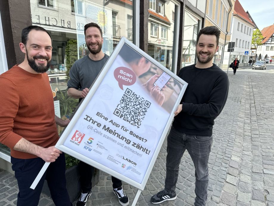 Stephan Siegert und Helge Ernst von der Stadt Soest halten zusammen mit Dennis Wiosna von SWCode ein Plakat zur Teilnahme an der SoestApp-Umfrage hoch.