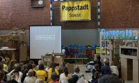 Das Foto zeigt eine große Gruppe von Kindern, die in einer Turnhalle sitzen und auf eine kleine Leinwand schauen. Sie sind von kleinen Gebäuden aus Pappe umgeben und schauen einen Film über die Pappstadt Soest. An der Rückwand der Halle hängt ein großes gelbes Ortsschild mit der Aufschrift Pappstadt.