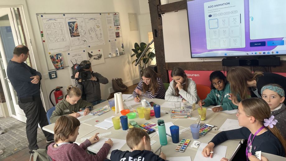 Kinder am Tisch, die malen und ein Fernsehteam im Hintergrund