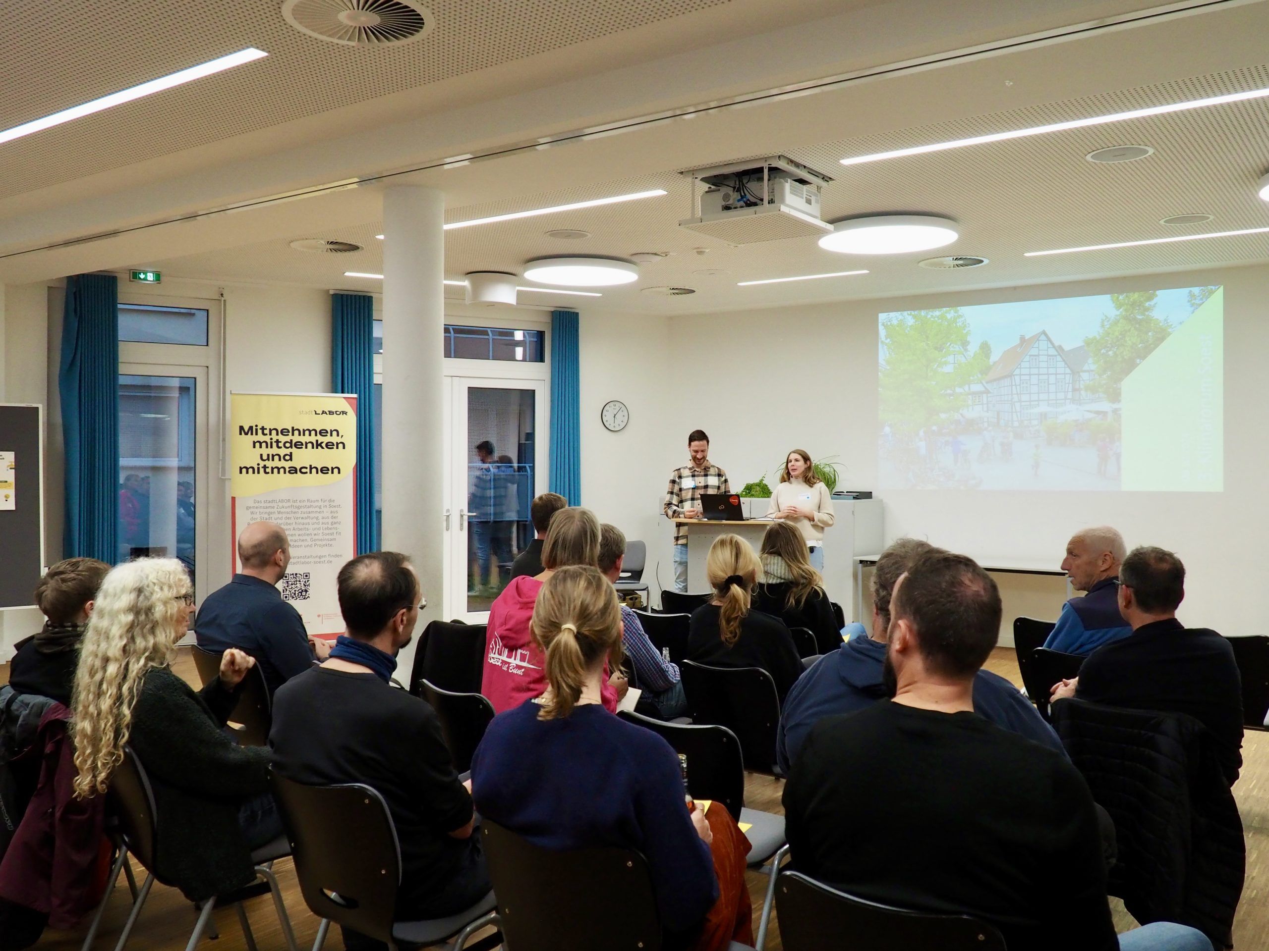 Ein Seminarraum, in dem vor einem Publikum von 20 Personen zwei Vortragenden die Abendveranstaltung Klimaforum moderieren. Im Hintergrund ist eine Popwerpoint-Präsentation an der Wand zu sehen. Die Vortragenden gestikulieren und Blicken in die Menge.