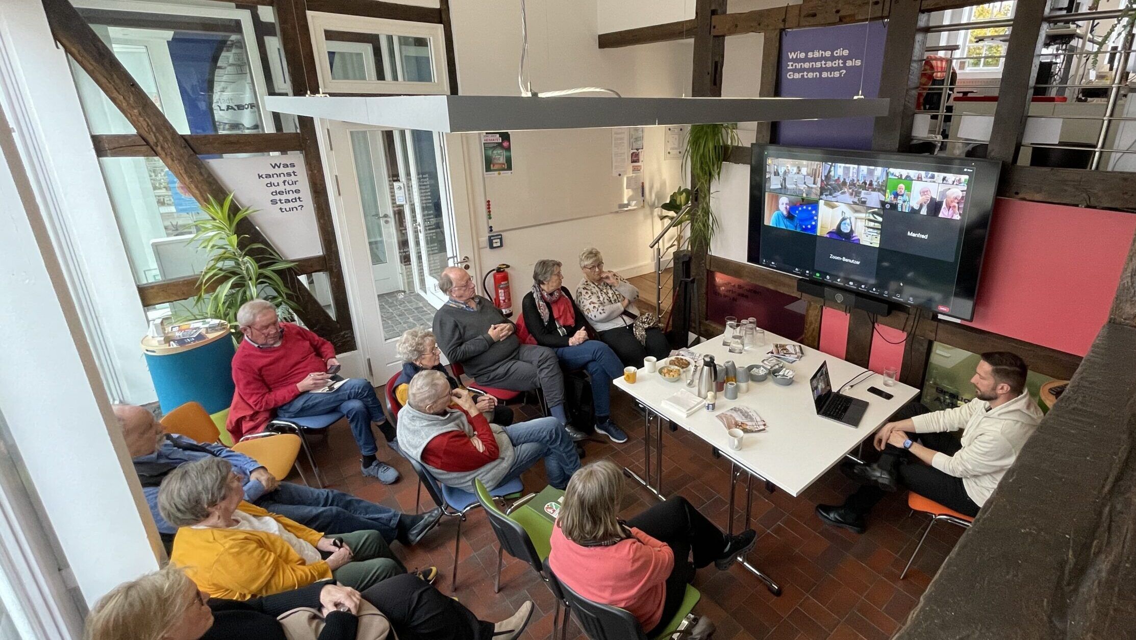 Das Bild zeigt eine Gruppe von älteren Menschen im stadtLABOR Soest, die interessiert einem Online-Vortrag zuhören.