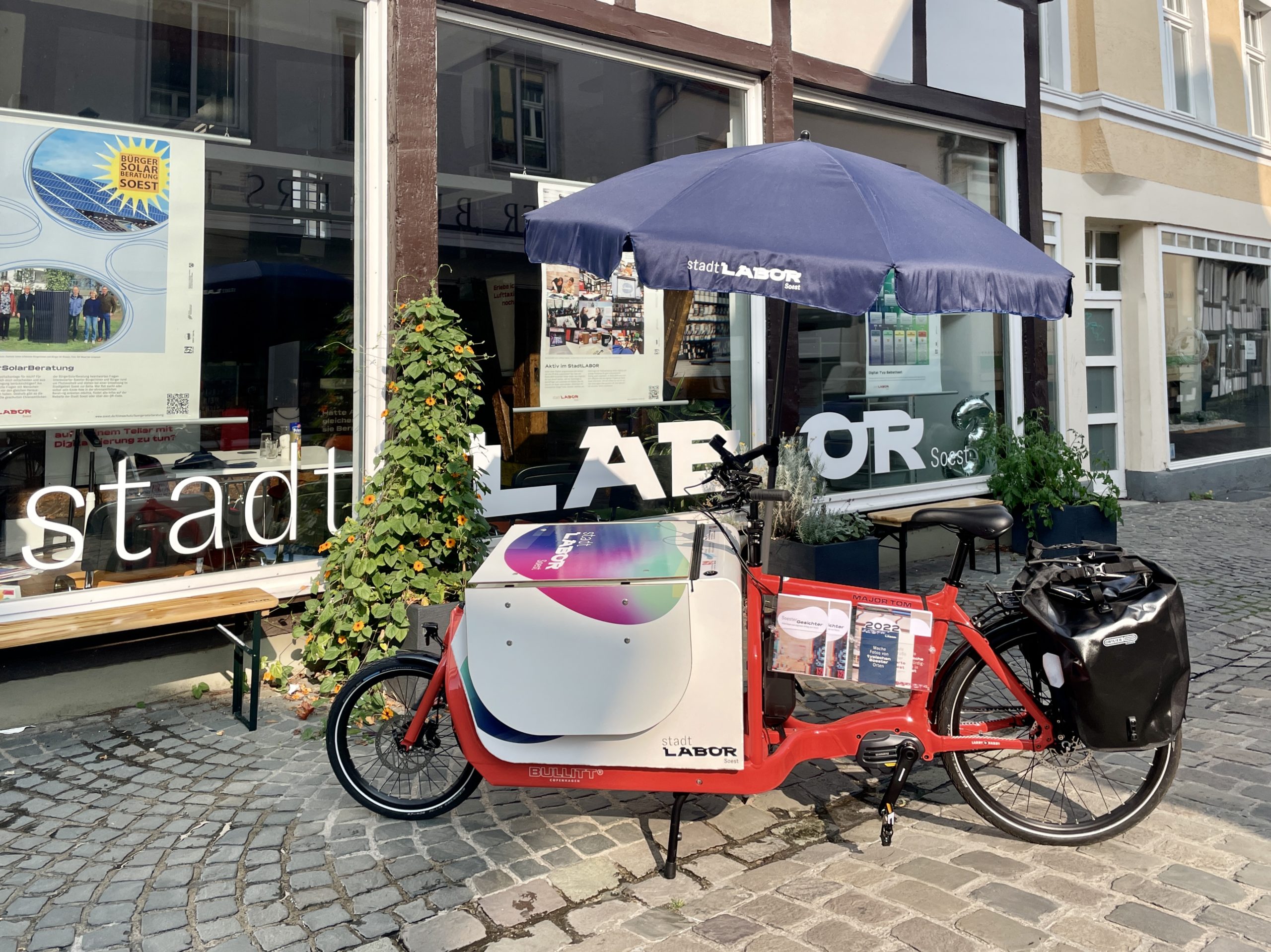 Ein Lastenrad mit Sonnenschirm und Tisch steht vor der sonnenbeschienen Front des stadtLABORs Soest.