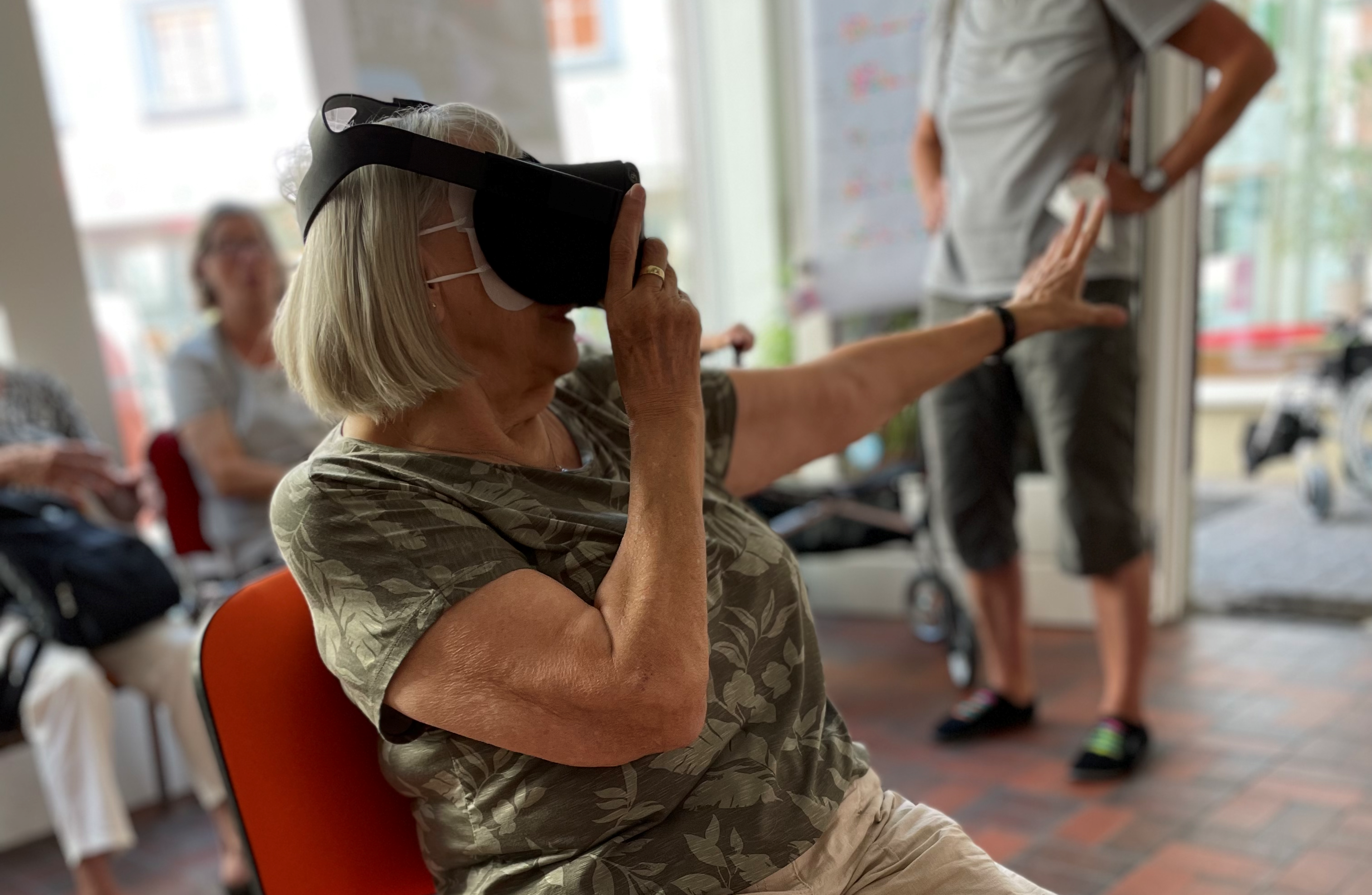 Eine ältere Dame sitzt auf einem Stuhl und trägt eine VR-Brille. Sie greift in die Luft, so als könnte sie dort etwas aus der virtuellen Welt anfassen.