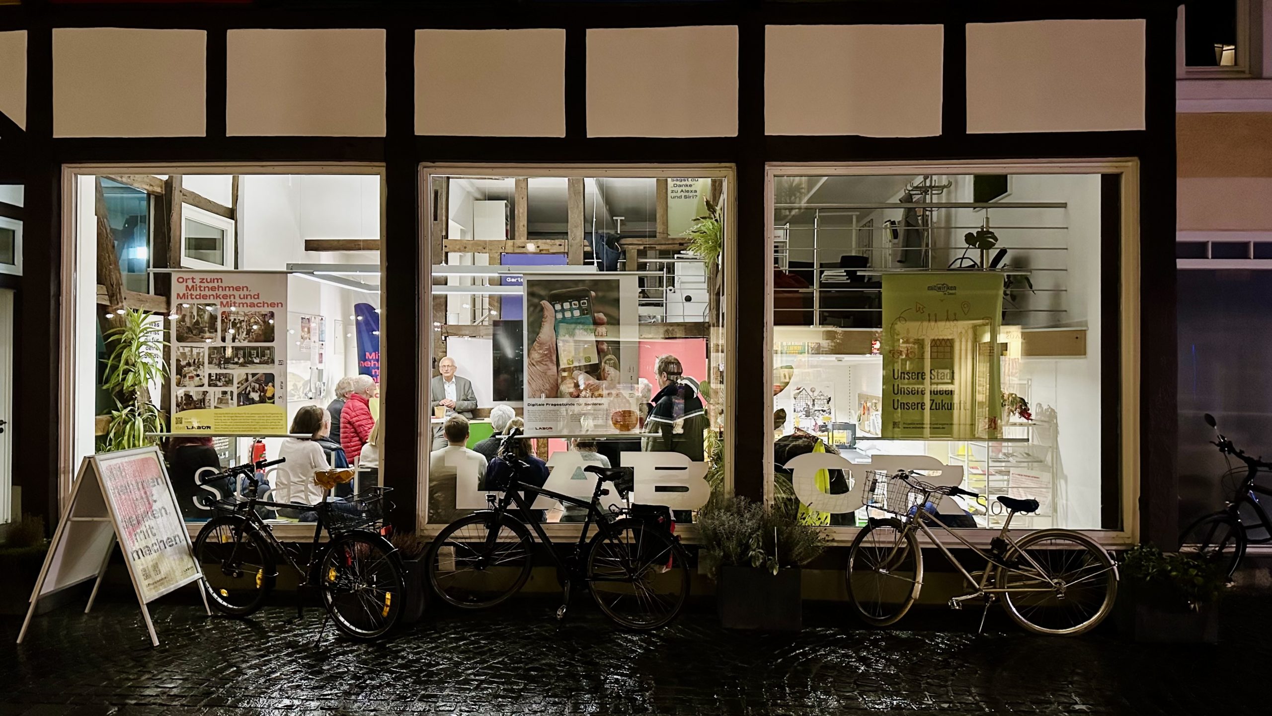 Das Bild zeigt das stadtLABOR Soest von außen. Es ist bereits Abend, aber durch die großen bodentiefen Fenster scheint Licht auf die Straße und reflektiert sich in auf dem regennassen Boden. Drinnen sind viele Menschen zu sehen, die in eine Diskussion vertieft sind.