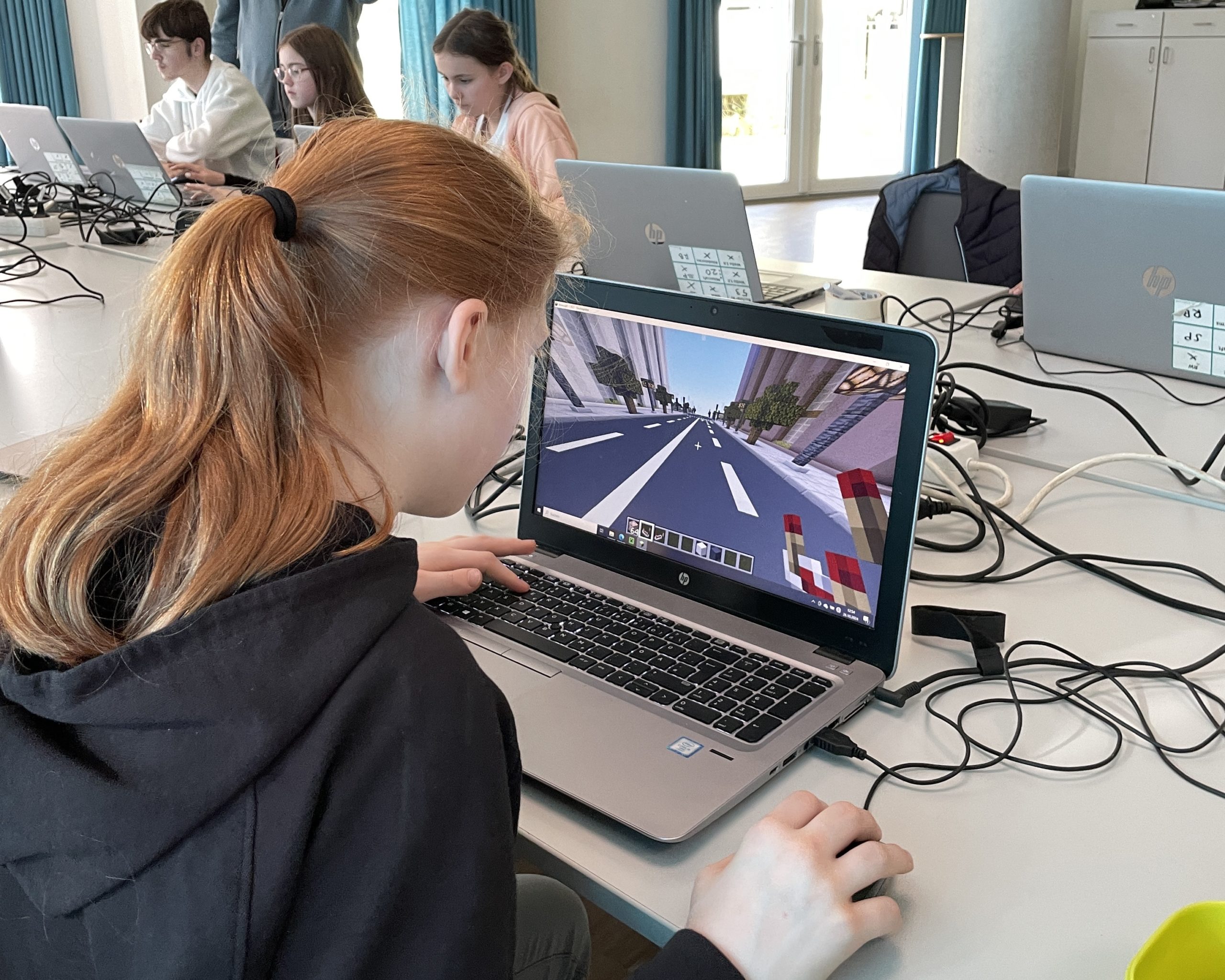 Ein Mädchen sitzt an einem Laptop und programmiert bei Minecraft. Im Hintergrund sind weitere Kinder an Laptops zu sehen.