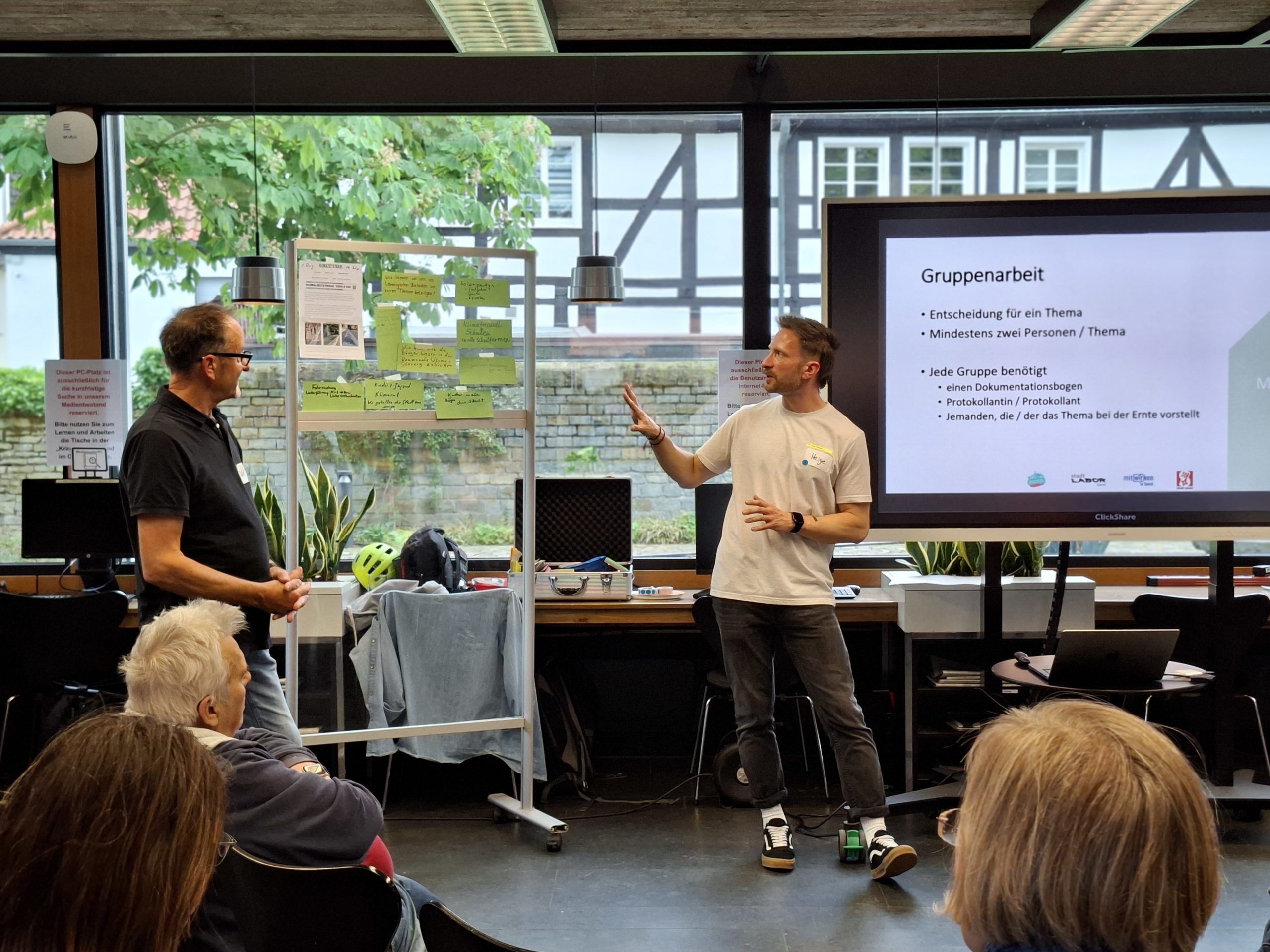 Menschen sitzen bei der Veranstaltung Klimaforum und hören zwei Mitarbeitenden der Stadt zu, die vorne stehen und etwas präsentieren.