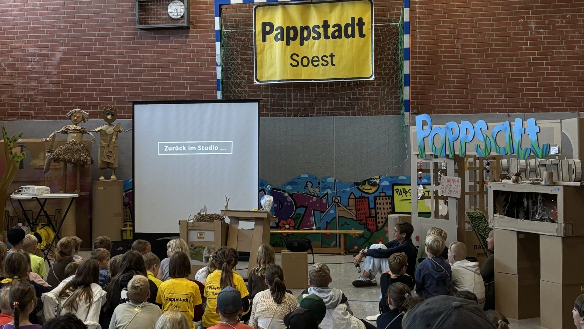 Das Foto zeigt eine große Gruppe von Kindern, die in einer Turnhalle sitzen und auf eine kleine Leinwand schauen. Sie sind von kleinen Gebäuden aus Pappe umgeben und schauen einen Film über die Pappstadt Soest. An der Rückwand der Halle hängt ein großes gelbes Ortsschild mit der Aufschrift Pappstadt.