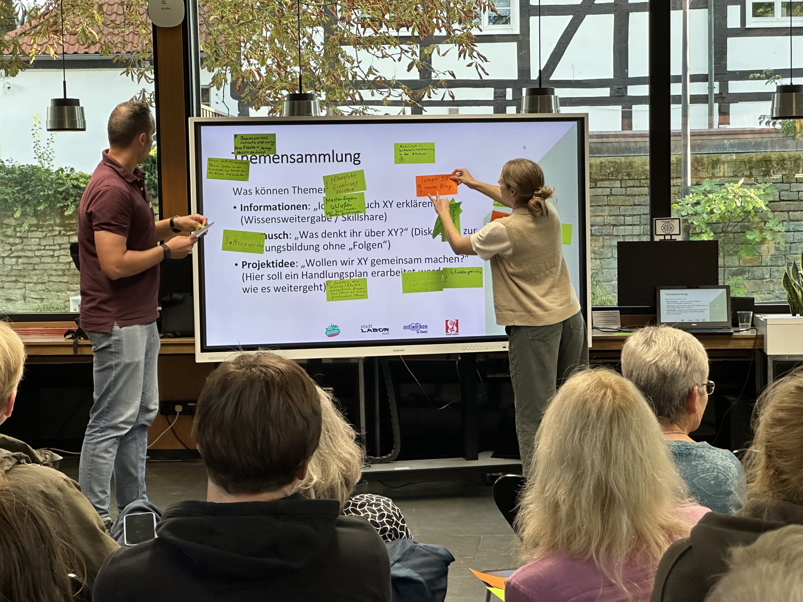 Eine Frau und ein Mann stehen vor einer Gruppe und kleben Kärtchen an ein Whiteboard