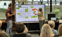 Eine Frau und ein Mann stehen vor einer Gruppe und kleben Kärtchen an ein Whiteboard