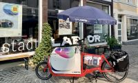 Ein Lastenrad mit Sonnenschirm und Tisch steht vor der sonnenbeschienen Front des stadtLABORs Soest.